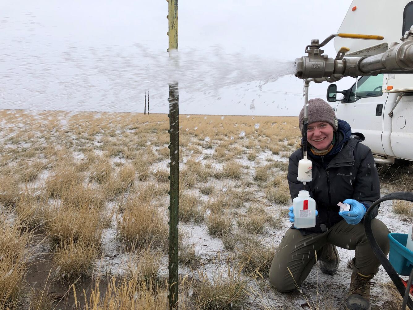 A person wearing a winter jacket collecting a groundwater sample from a pipe in a field.