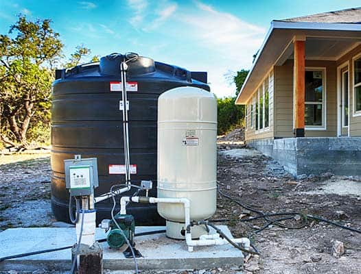 A black water storage tank and a beige pressure tank with an attached control panel and pump system are installed outside a house.