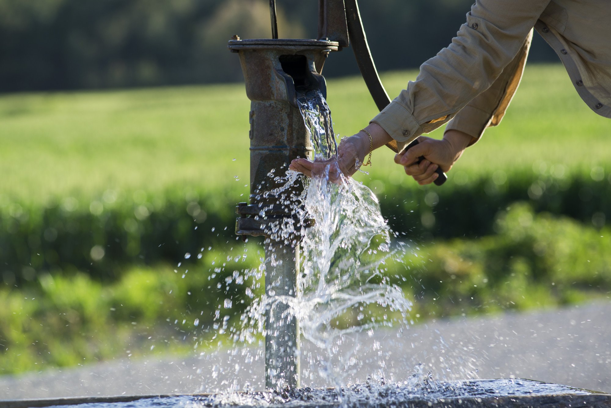 A person is pumping water from a hand pump outdoors, with water splashing as they catch it in their hands.