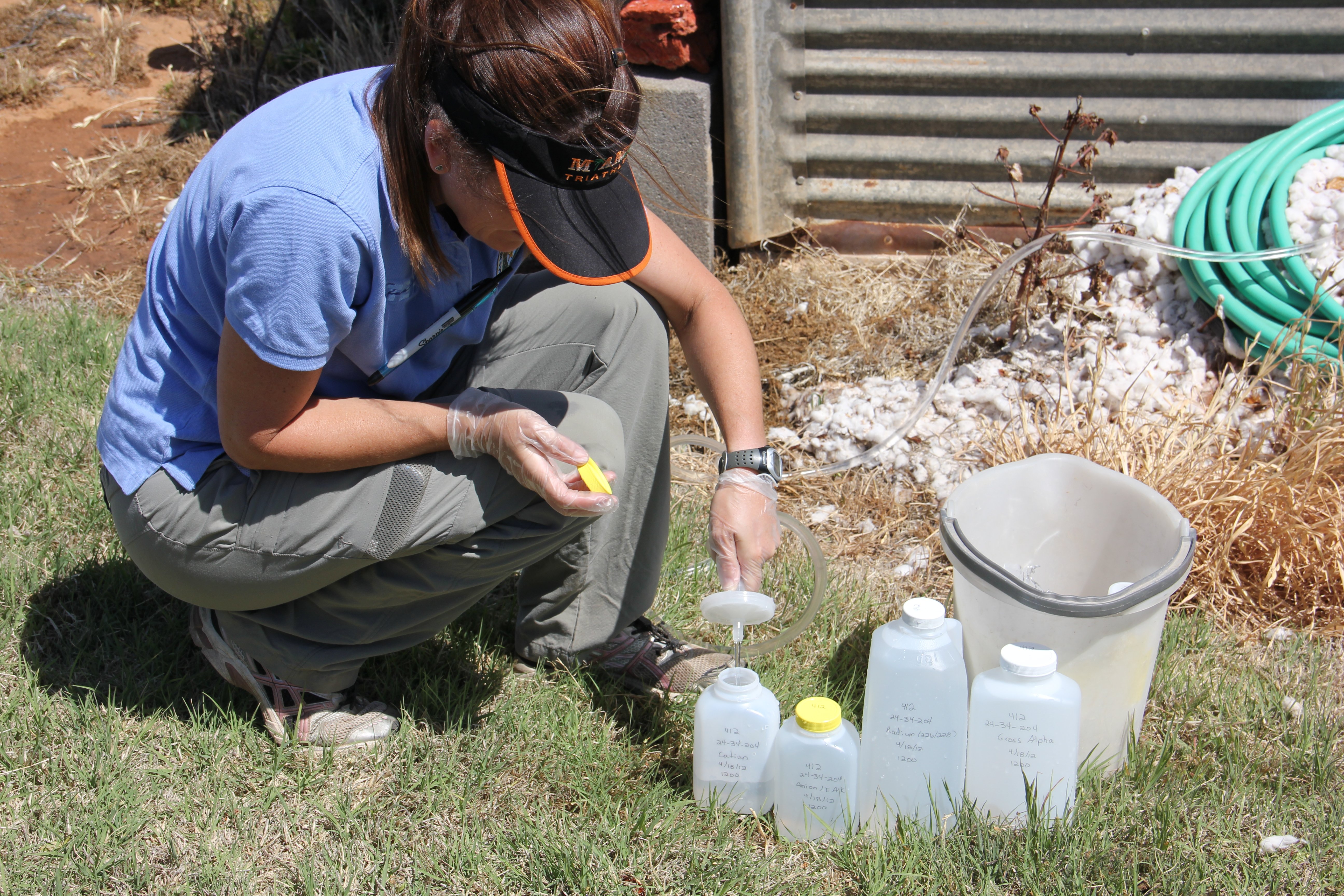 A person kneeling on grass is conducting a water sampling test using several bottles and equipment near a garden area with hoses and a bucket.