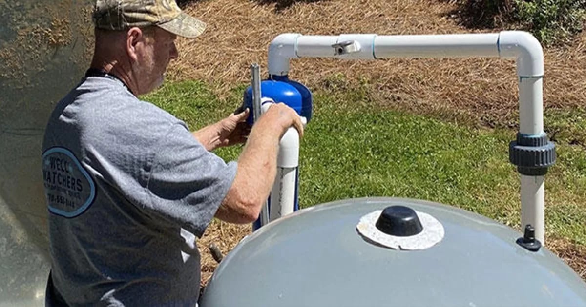 A person wearing a cap and a grey T-shirt is working on a pipeline setup in an outdoor setting with grass and mulch in the background.