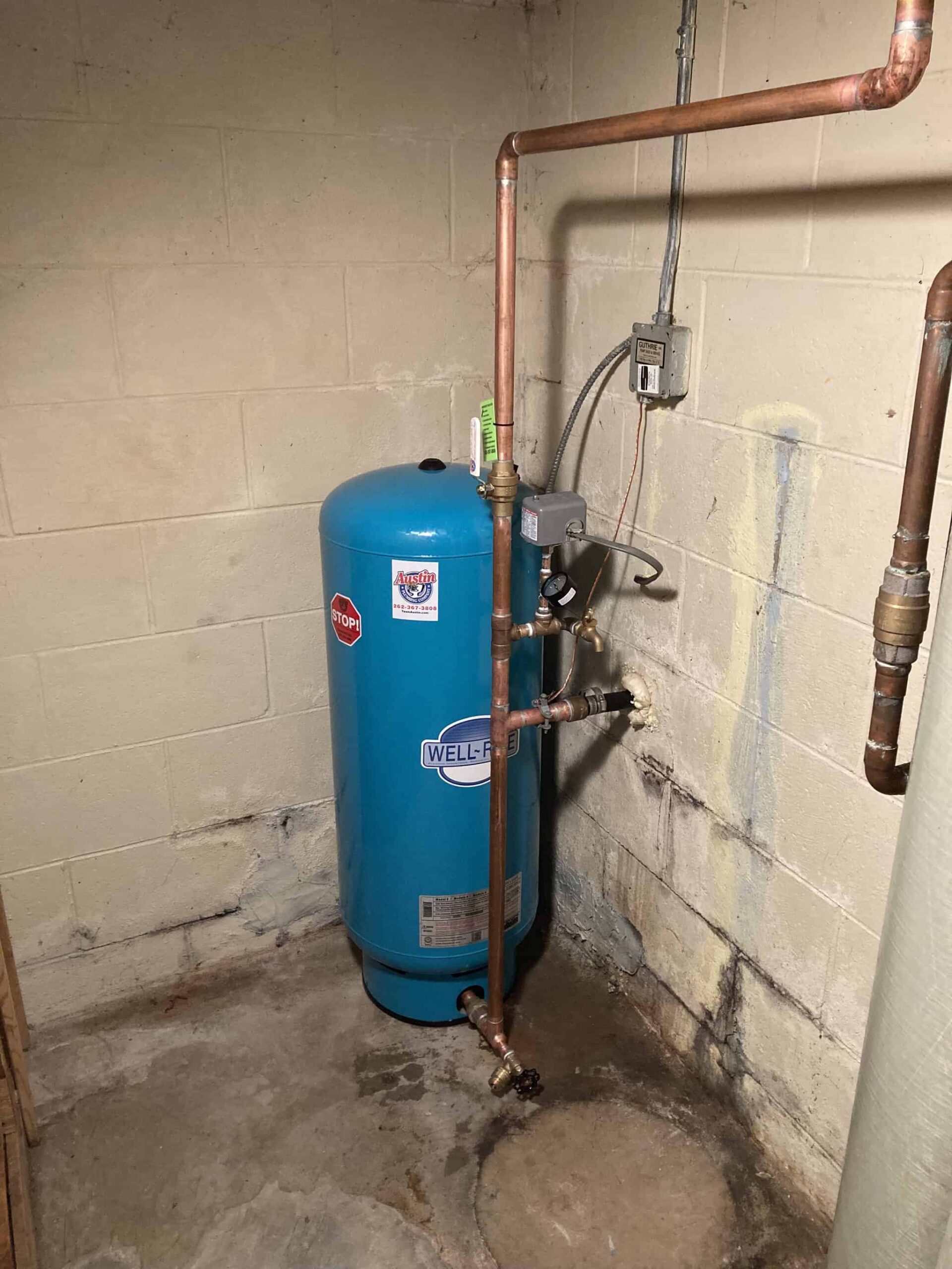 A blue Well-Rite pressure tank with attached copper pipes is installed in a basement with concrete walls and floor.