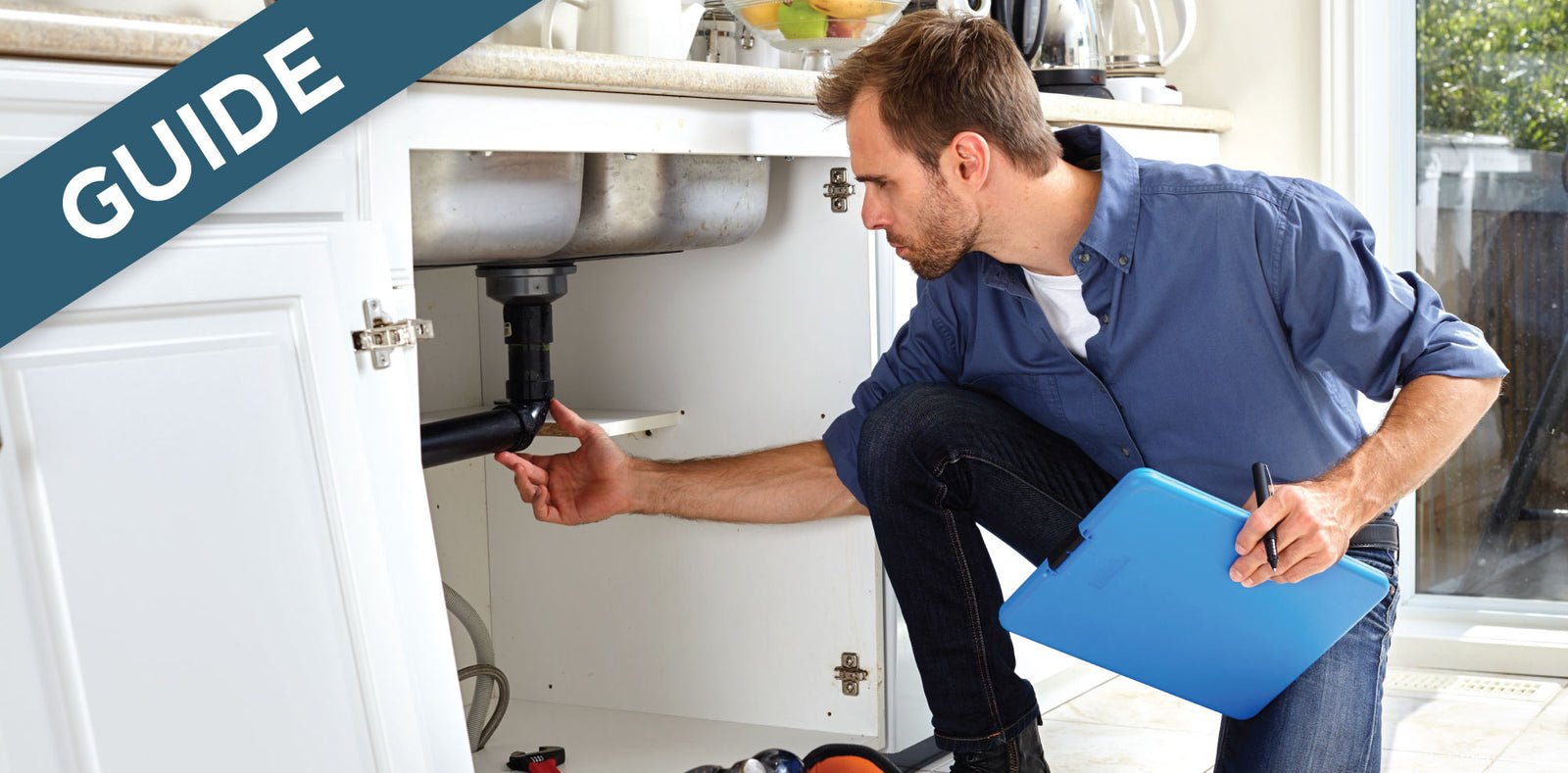 A person in a blue shirt inspects the plumbing under a kitchen sink, holding a blue clipboard and pen.